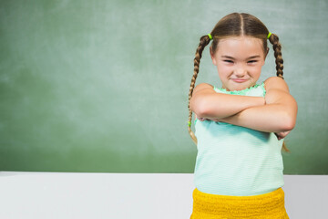 Female child standing with crossed arms in classroom at green chalkboard by white desk, copy space