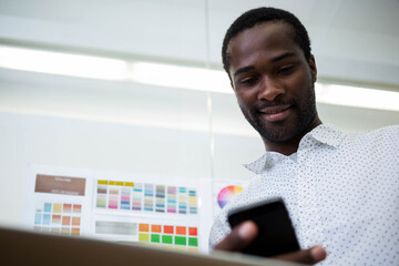 African American man standing in office checking phone beside laptop and color charts, copy space