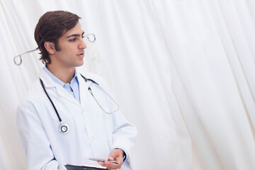 Male physician standing beside curtain and iv pole in clinic wearing stethoscope, holding clipboard
