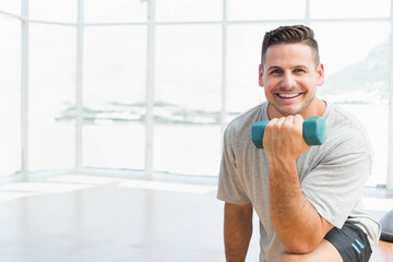 Man kneeling on gym floor wearing gray t-shirt holding teal dumbbell in bright studio, copy space