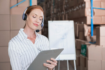 Woman standing in warehouse using headset writing clipboard notes near flip chart, copy space