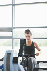 Woman pulling rower handle while monitoring digital screen in gym by windows in sportswear