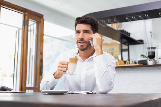 Middle-aged man in shirt talking on smartphone and holding latte glass at bright café table