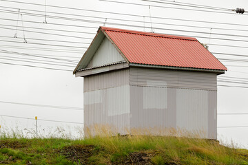 Small corrugated metal utility building with a red roof located on grassy terrain under multiple...