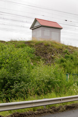 A small shed with a red roof stands on a green embankment near railway wires, surrounded by...