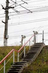 Outdoor concrete stairs with red handrail ascend a grassy slope toward electrical infrastructure...