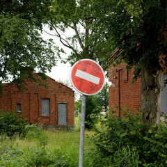A weathered no entry traffic sign stands beside overgrown grass and red brick buildings in a quiet,...