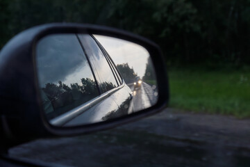 View of a car's side mirror showing a dark wet road with approaching headlights, trees, and...