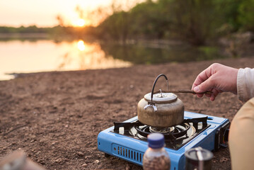 Person heating water in a kettle with a travel stove on the bank of a stream, at sunrise, to make coffee. El Palmar National Park, Entre Rios, Argentina.