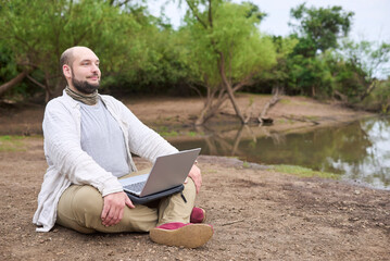 Hispanic man sitting outdoors, smiling while working on his laptop. Concept: digital nomad working remotely while traveling, enjoying a lifestyle of freedom and contact with nature.