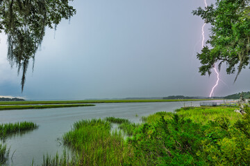 Lightning strikes over a calm river surrounded by lush green vegetation during a stormy evening in...