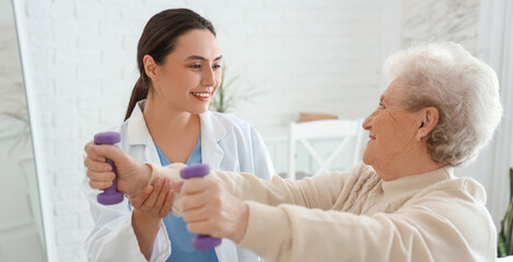 Fototapeta premium Physical therapist and senior woman training with dumbbells in bedroom