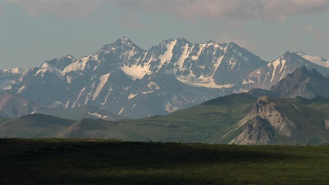 Snow covered peaks with rugged rocky ridges and green foothills in Denali National Park, Alaska. Static daytime view.