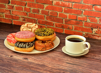 A cup of hot black coffee and a large plate with various fresh donuts in multi-colored glaze on a wooden table.