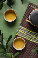 Bamboo mat with cups of tasty tea and teapot on green background