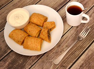Freshly baked pancakes stuffed with meat and sour cream on a wooden table, a mug of hot tea, a fork and a cup of sour cream nearby.