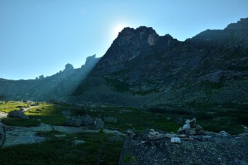 The rising sun casts bright rays on the edge of a high cliff casting a shadow on a picturesque mountain valley on a sunny summer morning.