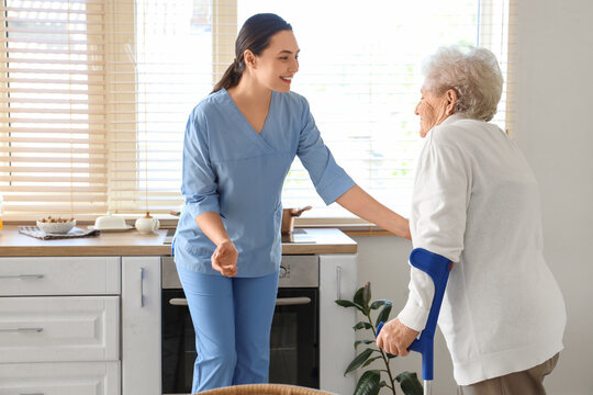 Physical therapist helping senior woman with crutches in kitchen - Powered by Adobe