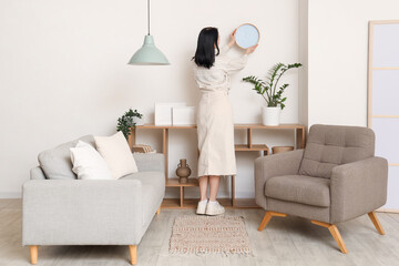 Young woman hanging clock near shelving unit in room. Back view