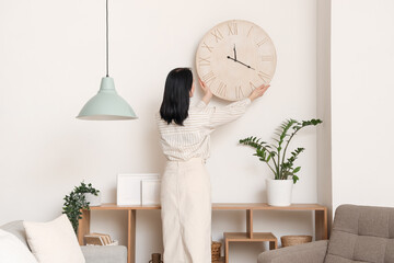 Young woman hanging clock near shelving unit in room. Back view