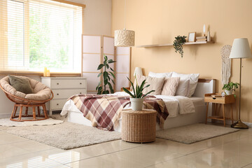 Interior of light bedroom with bed, armchair, table and folding screen near beige wall
