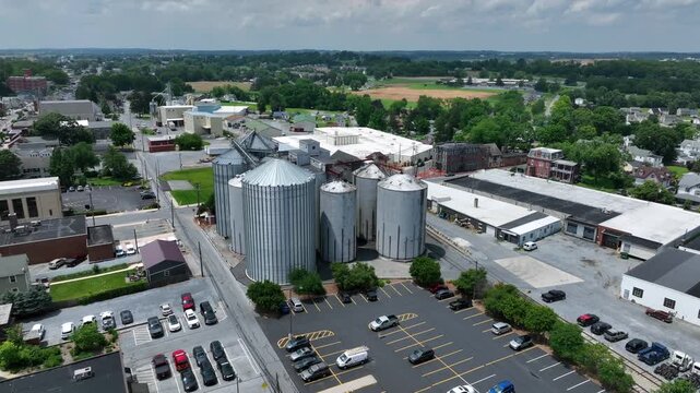 Grain silo tanks in city of America. Aerial approaching shot. Parking cars of workers in front of factory. Sunny day in Pennsylvania, USA.