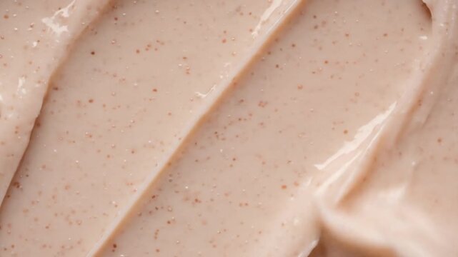 Closeup of light pink scrub or cream with small brown exfoliating beads spread in diagonal strokes reflecting light