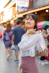Young Asian woman carrying a plastic bag of snacks on a bustling street during golden hour, with...