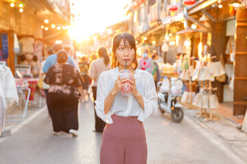 Young Asian woman enjoying a refreshing drink with a straw during sunset at a lively outdoor street...