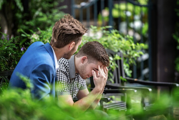 A man is sitting on a bench with his head in his hands. Another man is sitting next to him,...
