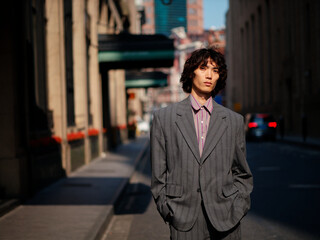 Portrait of handsome Chinese young man wearing gray suit posing in the street, young guy with black curly hair with urban background. Male fashion, cool Asian young man lifestyle.