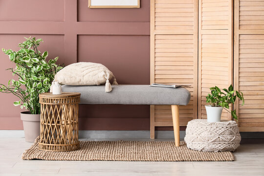 Soft bench, folding screen, table and pouf near brown wall in interior of living room, closeup