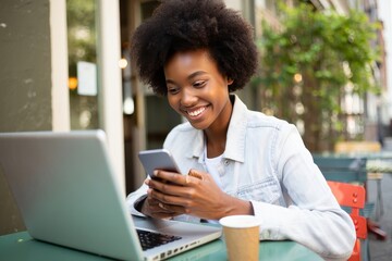 Smiling Black Woman Using Smartphone at Outdoor Cafe with Laptop