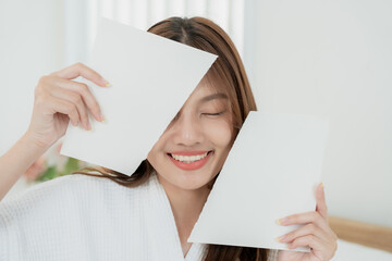 Asian teenage girl in white robe holding two blank white sheets near face while smiling with eyes...