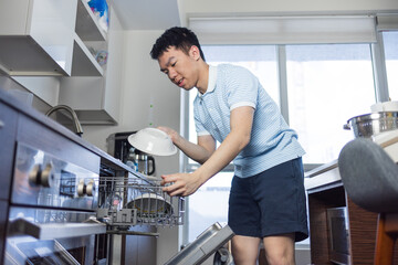 A young man in casual clothes places dishes into an open dishwasher. Useful for topics on chores,...