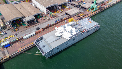 A modern military ship docked at an industrial shipyard, featuring advanced radar domes and a...