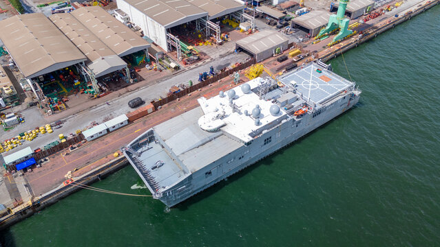 Fototapeta A modern military ship docked at an industrial shipyard, featuring advanced radar domes and a helicopter landing pad, surrounded by cranes, warehouses, and marine equipment.