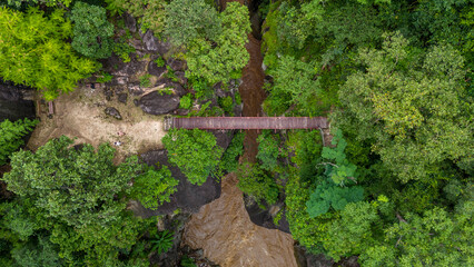 Aerial view of a wooden footbridge crossing a muddy river surrounded by dense green forest and rocky terrain, highlighting nature, adventure, and eco-tourism