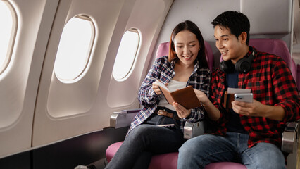A young Asian couple sitting on an airplane, smiling while holding passports and a smartphone. They appear excited and ready for their journey, dressed in casual travel outfits.