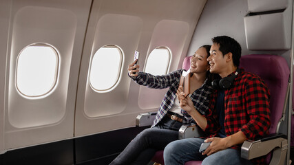 A young Asian couple sitting on an airplane, smiling while holding passports and a smartphone. They appear excited and ready for their journey, dressed in casual travel outfits.