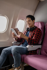 Asian man sitting comfortably on airplane seat near window, holding smartphone and passport, wearing headphones around neck, enjoying in-flight entertainment. Modern travel lifestyle and technology.