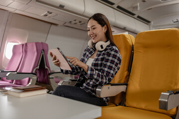 Young Asian woman sitting in airplane seat with yellow cover, smiling while using tablet during flight. She wears casual outfit with headphones around her neck, enjoying digital entertainment.