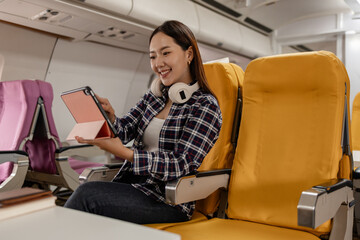Young Asian woman sitting in airplane seat with yellow cover, smiling while using tablet during flight. She wears casual outfit with headphones around her neck, enjoying digital entertainment.
