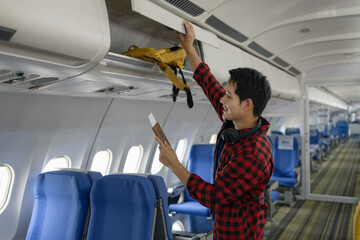Young Asian man placing a yellow backpack into an overhead compartment on an airplane. He looks focused while organizing his belongings before takeoff, dressed casually with headphones around his neck