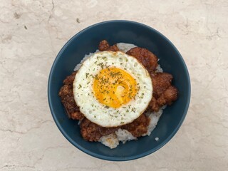 Close-up of rice bowl topped with fried chicken and sunny side up egg, garnished with herbs. Asian comfort food served in blue bowl on marble background.