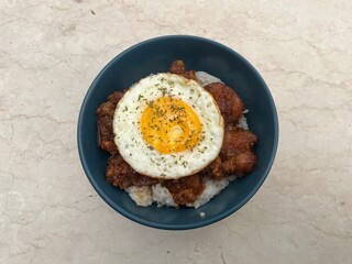 Close-up of rice bowl topped with fried chicken and sunny side up egg, garnished with herbs. Asian comfort food served in blue bowl on marble background.