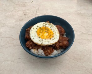 Close-up of rice bowl topped with fried chicken and sunny side up egg, garnished with herbs. Asian comfort food served in blue bowl on marble background.