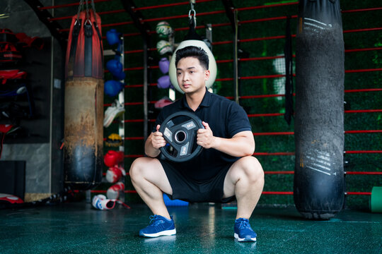 Young Asian man performing a squat exercise with a weight plate in a gym environment focusing on strength training and fitness