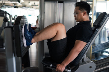 Young Asian man performs leg press exercise at gym with focused expression in bright, modern...
