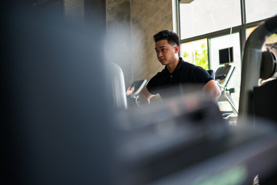 Young Asian man exercising in a modern gym setting during the morning, focused on his fitness routine while using an elliptical machine